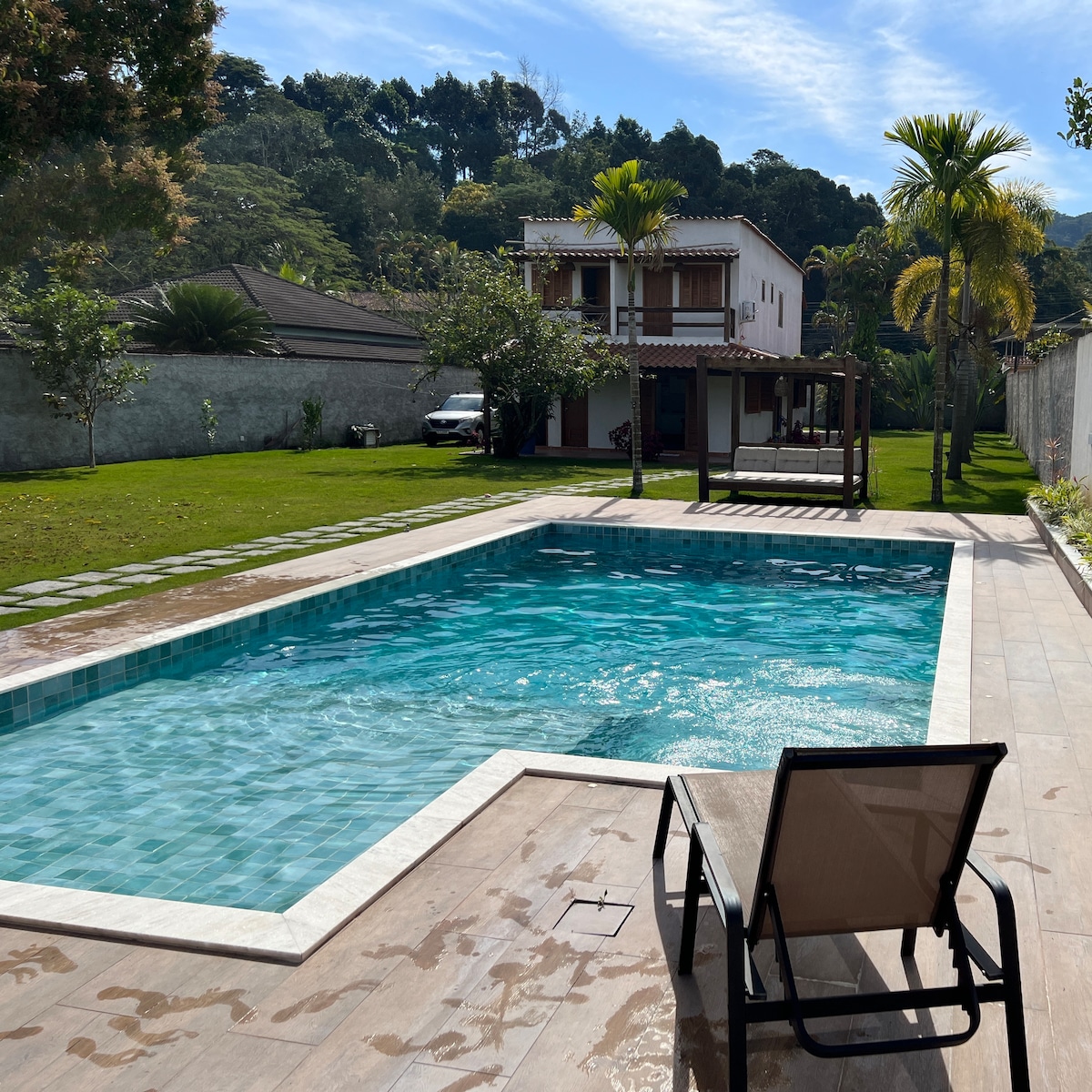 An inviting outdoor space features a spacious swimming pool with clear blue water, surrounded by a well-maintained lawn. A lounge chair sits at the pool's edge, and a wooden gazebo is visible in the background, alongside a modern two-story house framed by lush greenery.