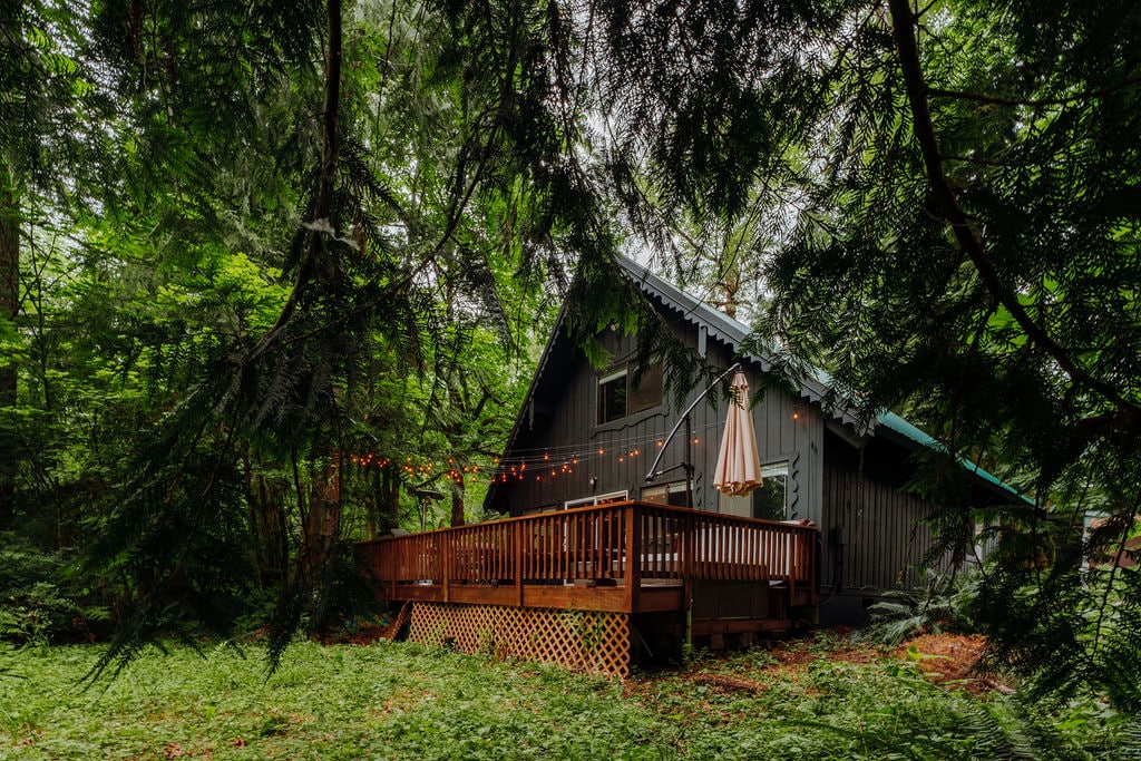 The cabin is nestled among lush greenery, featuring a spacious deck adorned with string lights and a large umbrella. The exterior showcases a pitched roof, blending harmoniously with the surrounding forest, providing a serene and inviting atmosphere.