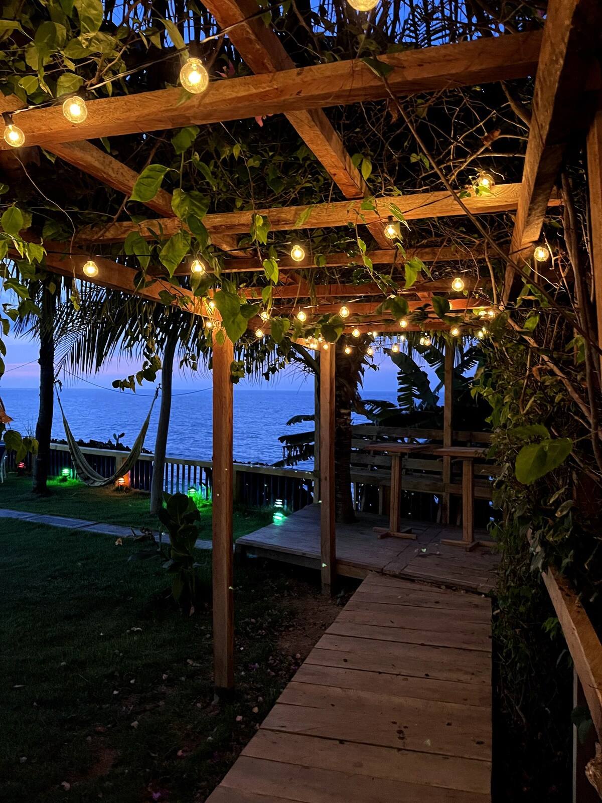 A wooden pathway leads through a welcoming outdoor area, lined with warm string lights and adorned with tropical plants. A hammock can be seen facing the ocean, providing a peaceful retreat, while the gentle blue hues of dusk create a serene atmosphere.