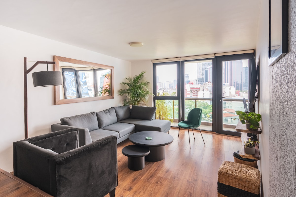 A modern living room features a comfortable gray sofa and an armchair, complemented by a round black coffee table. Large windows provide ample natural light and city views. A decorative mirror enhances the space, while a green plant adds a touch of nature.