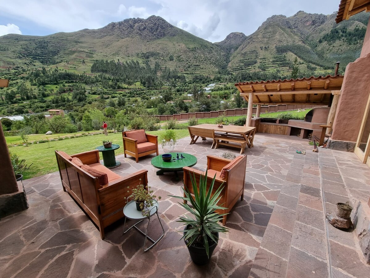 An outdoor seating area features wooden chairs and a low table, positioned on a stone patio. Lush green mountains serve as the backdrop, while a covered grill area is visible to the side, creating an inviting space for relaxation and gatherings.