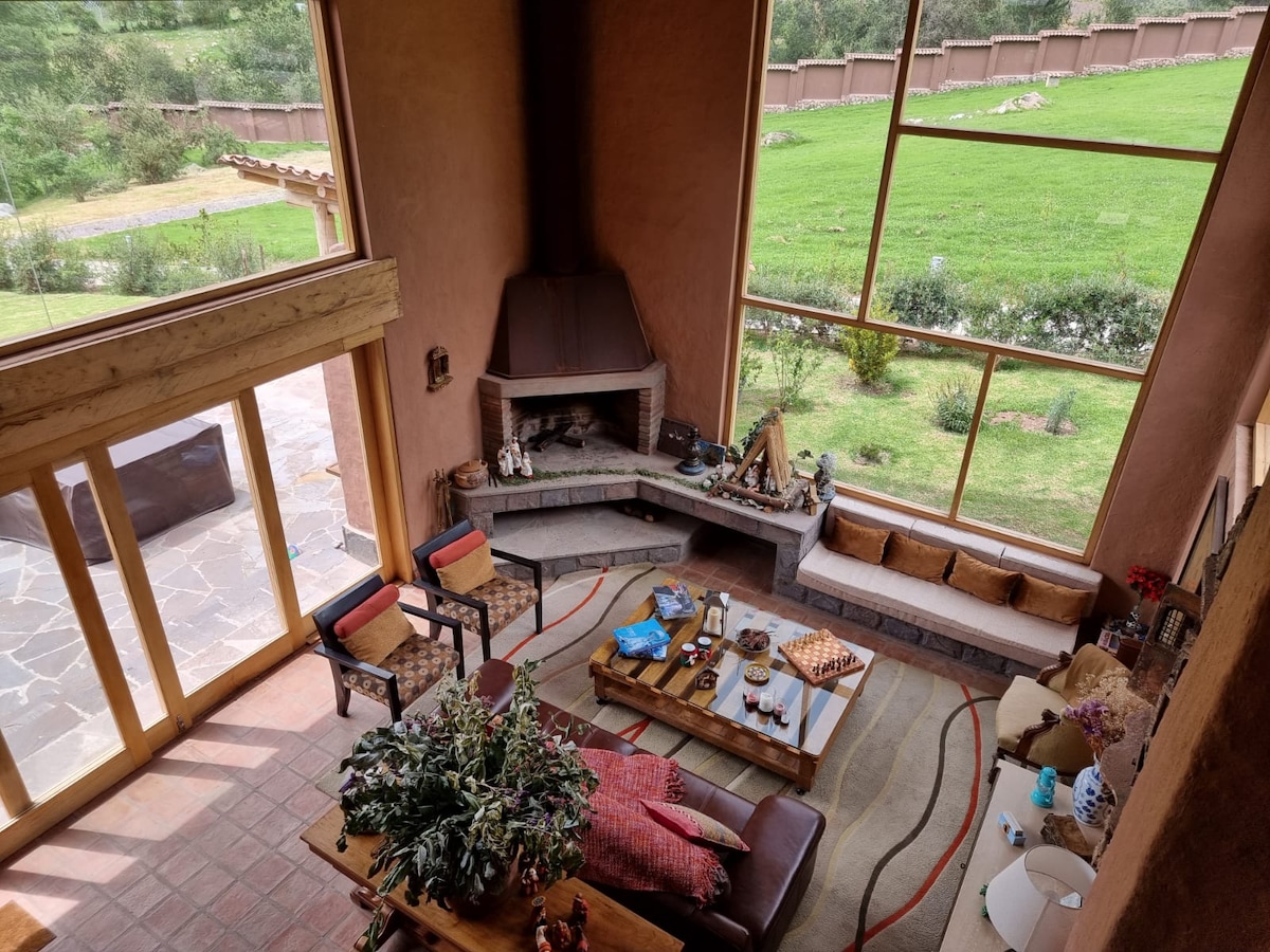 The spacious living room features a large corner fireplace and oversized windows, providing ample natural light and views of the green landscape outside. Comfortable seating areas are arranged around a wooden coffee table, with decorative plants and cozy cushions enhancing the inviting environment.