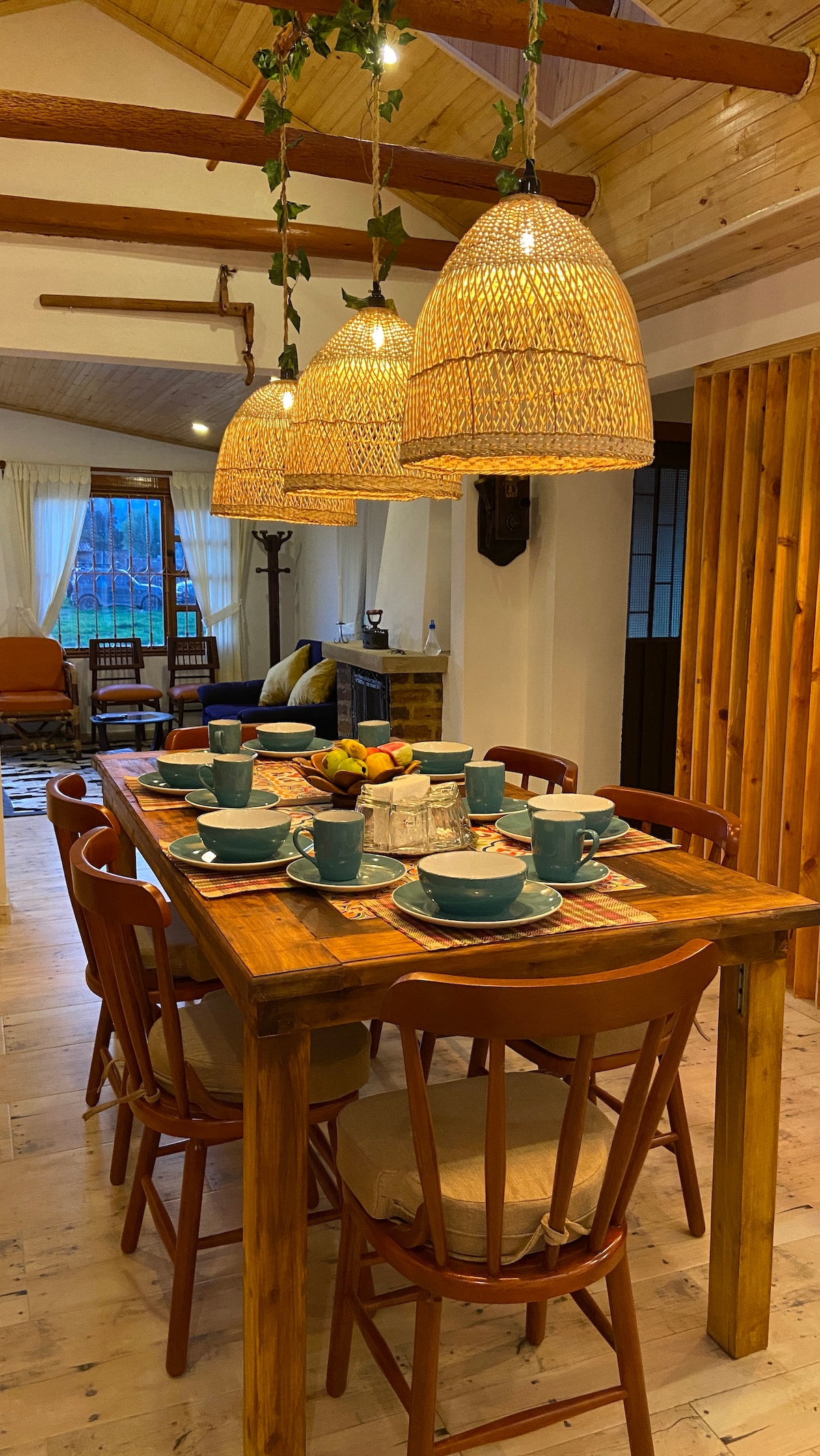 A wooden dining table is surrounded by six chairs, set for a meal with blue dishware. Three woven pendant lights hang overhead, illuminating the space. A cozy living area can be seen in the background, featuring a sofa and a fireplace.