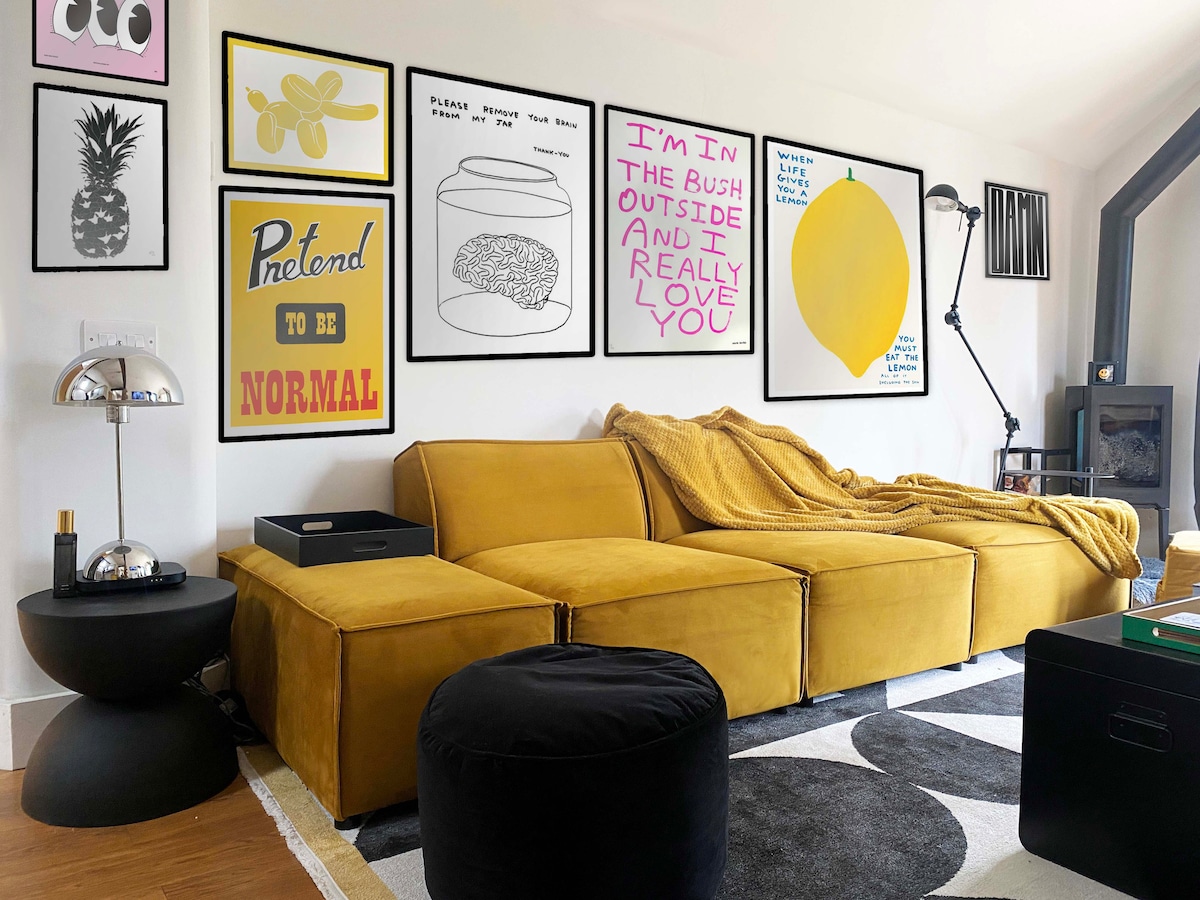 A vibrant living area features a spacious yellow sectional sofa with a cozy throw draped over it. The wall is adorned with various framed prints, adding a playful and modern touch. A plush black pouf and unique side tables anchor the seating arrangement in this inviting space.