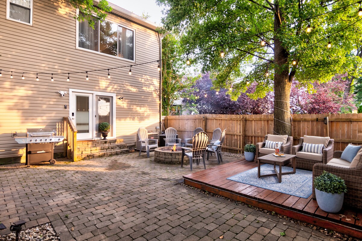 A spacious outdoor courtyard features paved stone flooring surrounded by a wooden fence. Seating areas comprise several lounge chairs and a fire pit, while a deck with a rug hosts a table and chairs. Soft lighting is provided by string lights overhead.