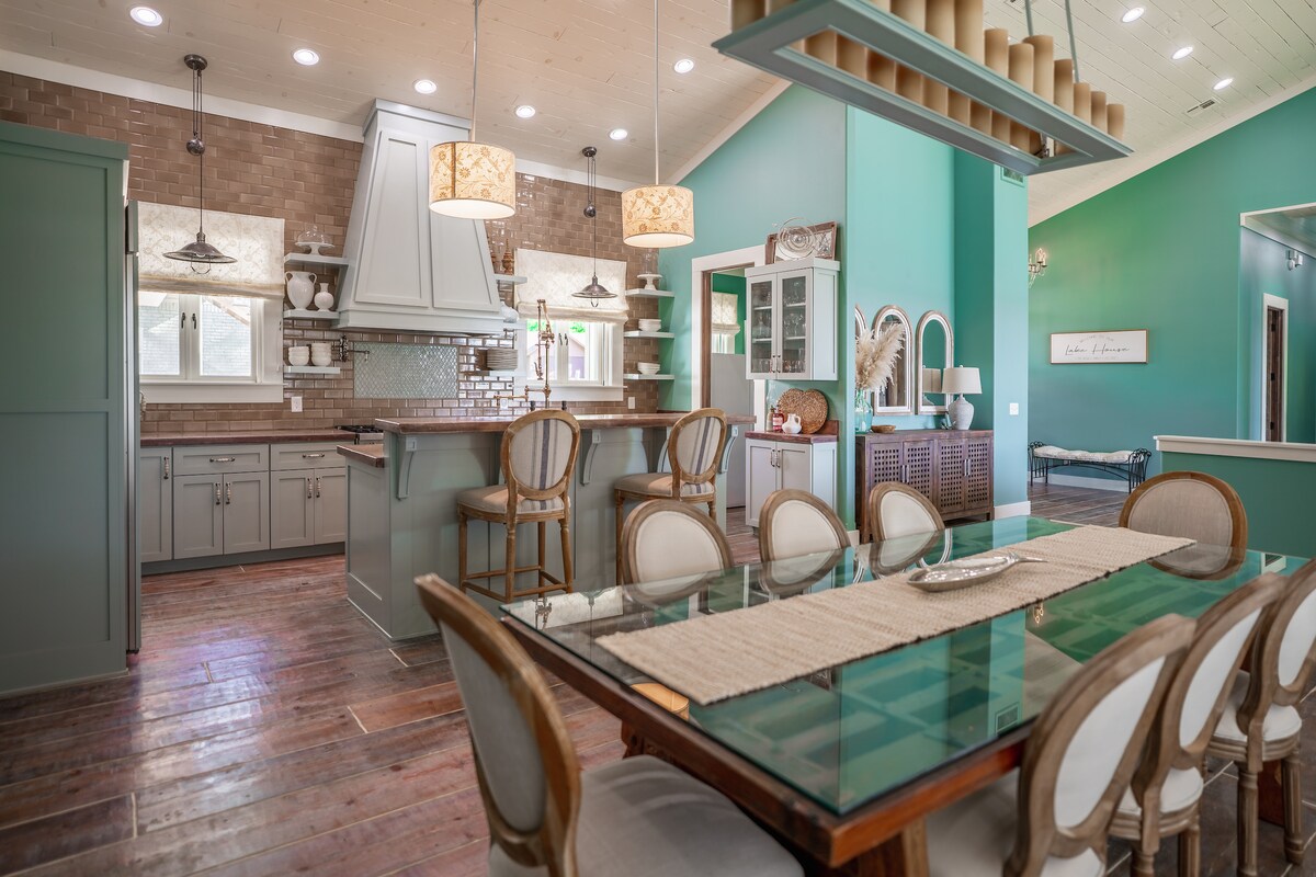 The dining area features a glass-top table surrounded by several upholstered chairs, illuminated by pendant lights. A modern kitchen is visible in the background, equipped with a large island, cabinetry, and warm-toned backsplash. Natural light fills the space through large windows.