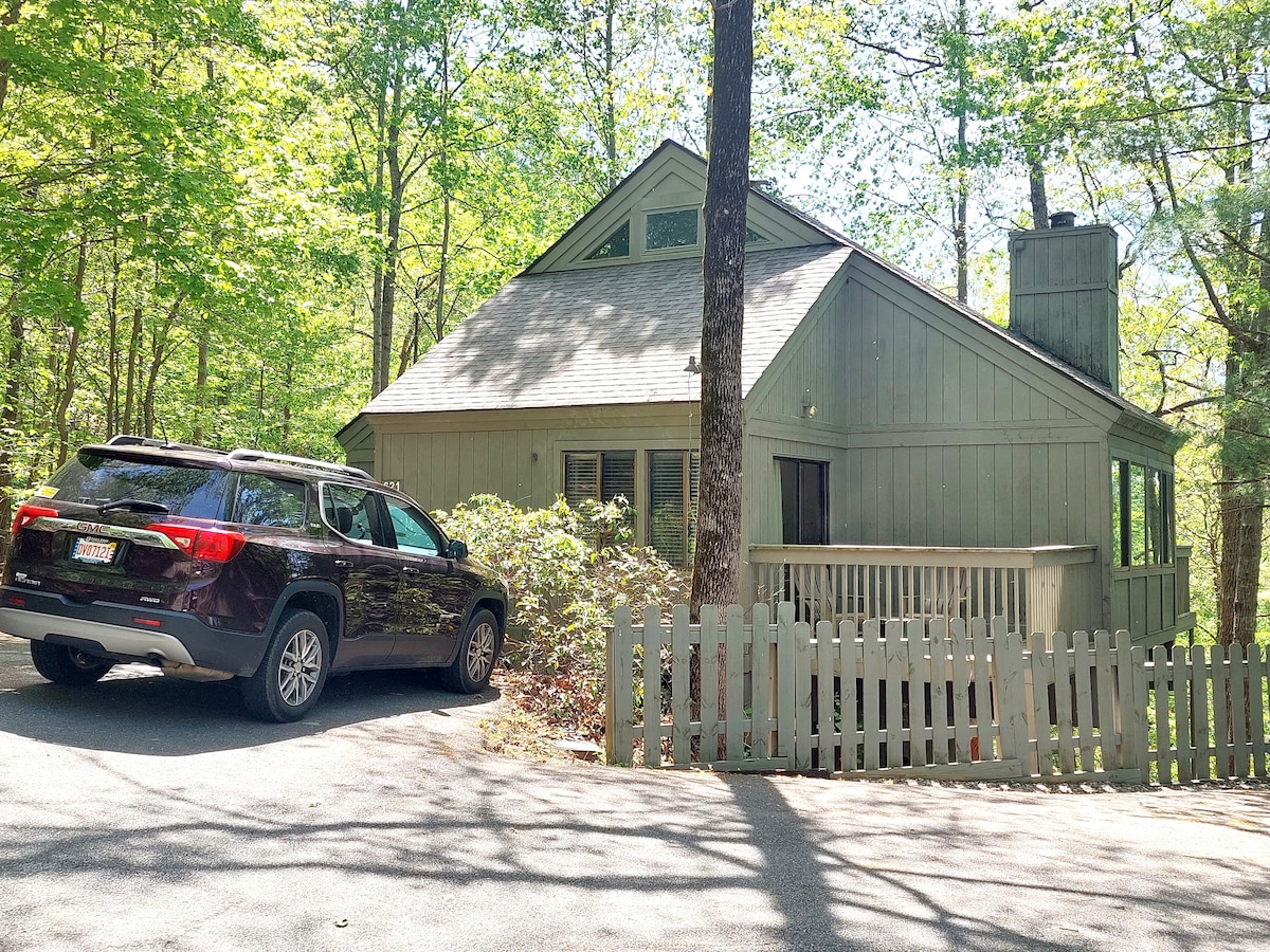 The exterior of a modern cabin is set amid lush greenery, featuring a sloped roof and large windows. A vehicle is parked in front, while a white picket fence lines the property, creating a welcoming entrance.