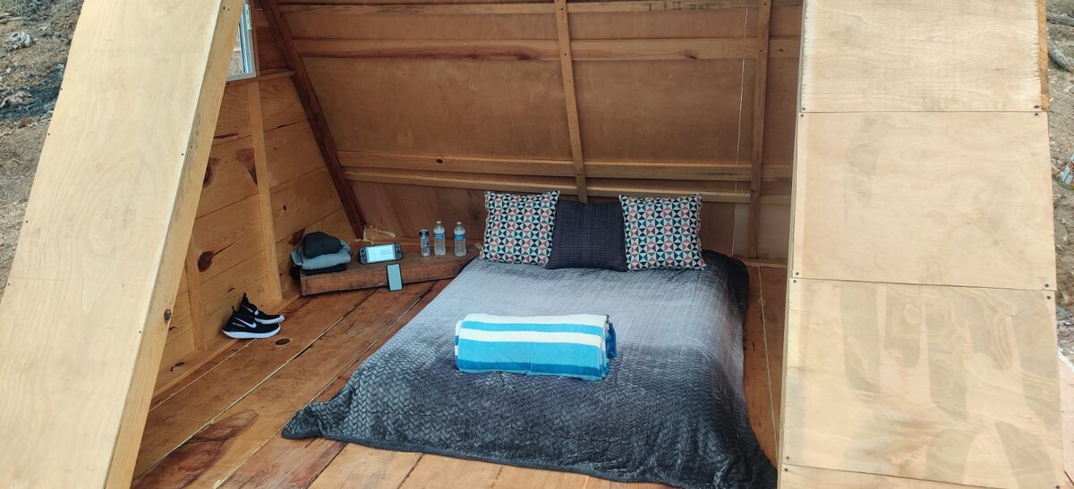 An inviting sleeping area features a spacious bed adorned with a soft gray blanket and colorful pillows. A rolled towel in blue is neatly placed at the foot of the bed. Surrounding elements include a wooden floor and a simple shelf with bottled water.