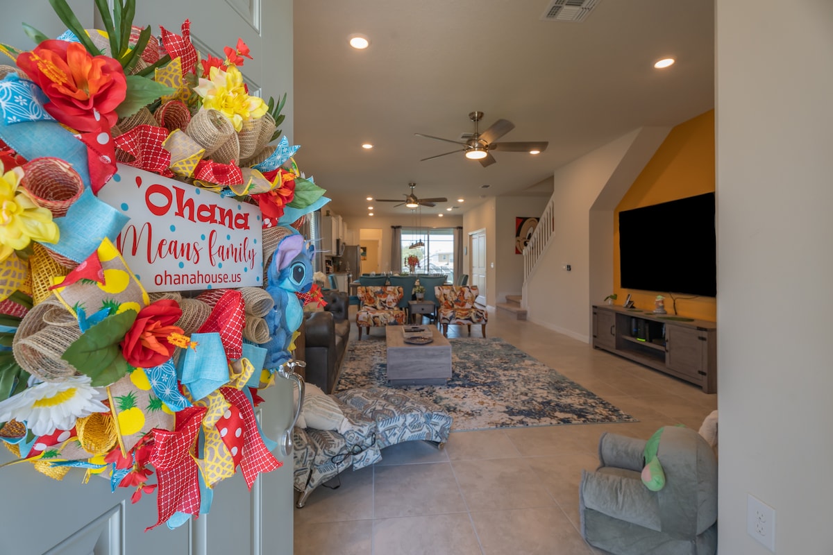 A welcoming entryway is adorned with a vibrant wreath featuring flowers and bright fabrics. Inside, a spacious living area is visible, furnished with comfortable seating and a large TV mounted on a feature wall. Natural light brightens the open space, enhancing the inviting ambiance.