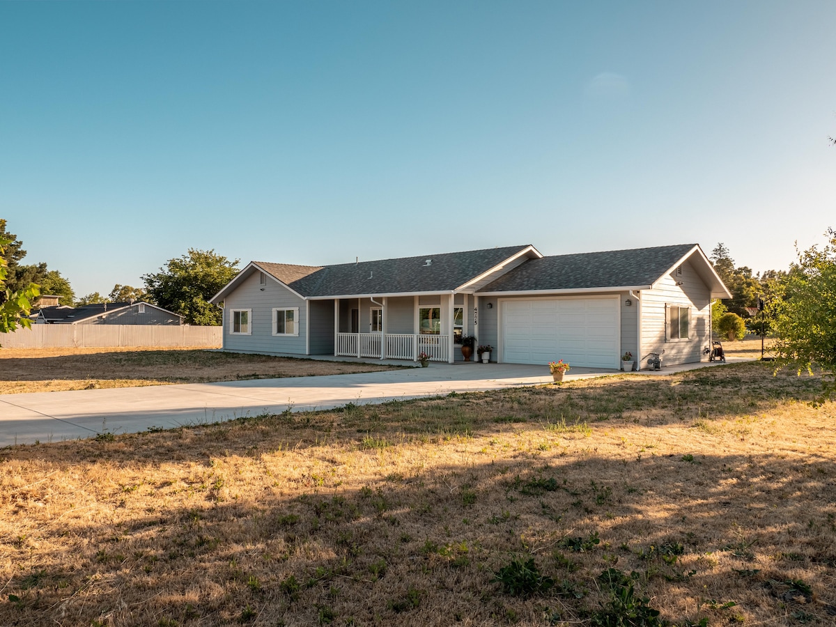 A spacious single-story home is showcased from the front, featuring a covered porch and a large driveway. The exterior is complemented by a manicured lawn and trees surrounding the property, with a fence visible in the background.