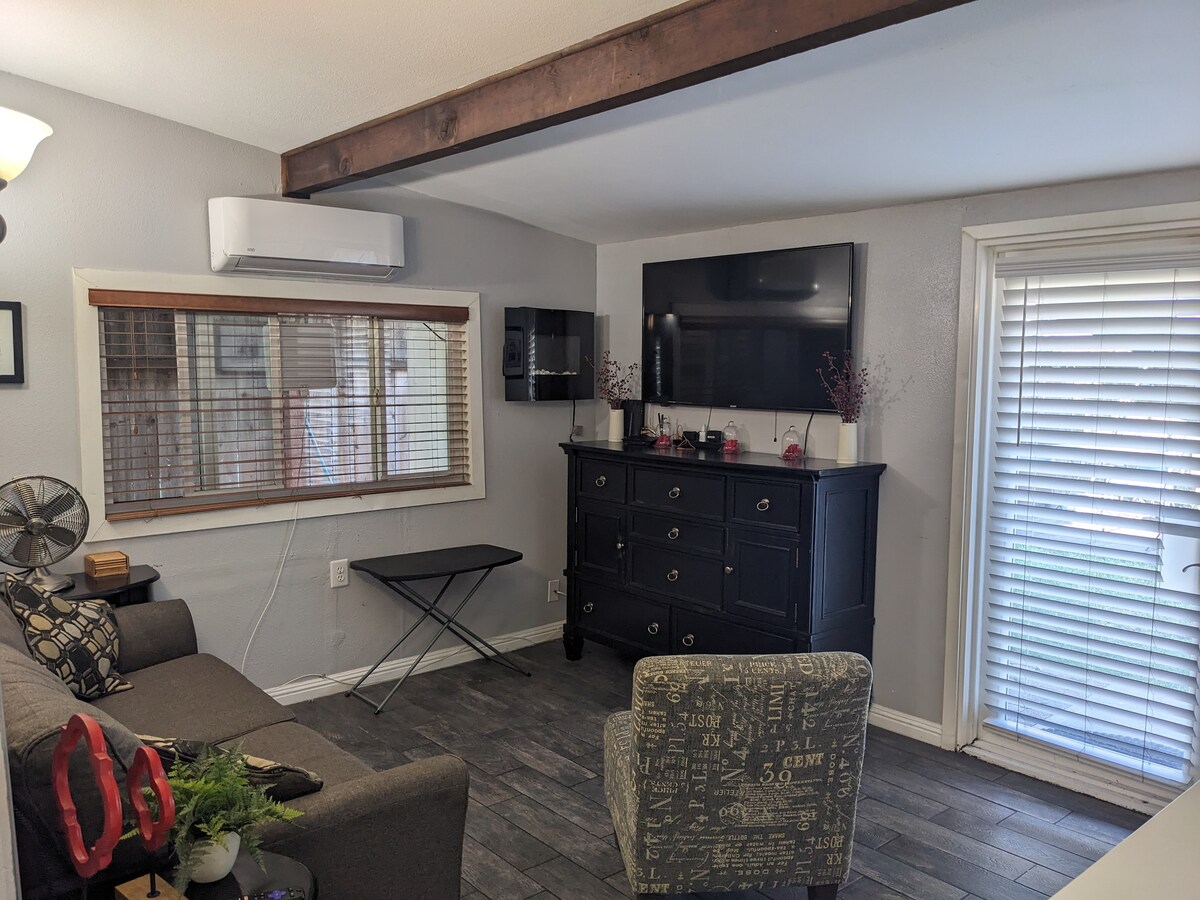 A living room area is highlighted by a mounted television and a dark wood dresser. Natural light enters through a window with blinds. A comfortable sofa and an accent chair provide seating, while a small folding table is positioned nearby. An air conditioning unit is mounted on the wall.