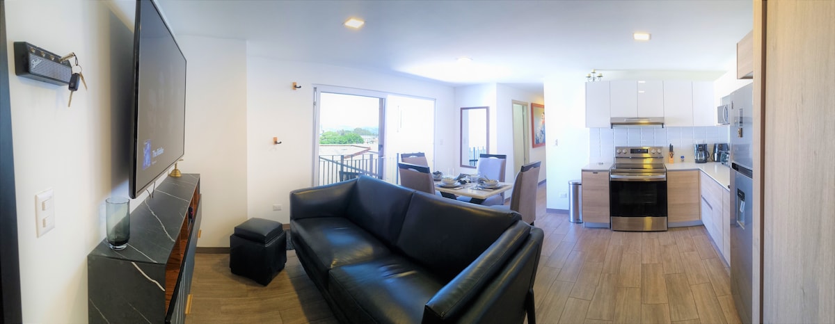 A spacious living area features a black leather sofa and a modern television mounted on the wall. To the right, a dining table is set for four, with a fully equipped kitchen visible in the background. Natural light flows through the large windows.