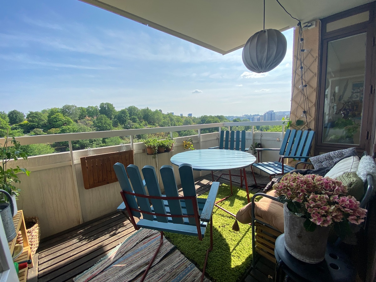 A spacious balcony is adorned with blue chairs and a round table, surrounded by greenery and potted plants. A rug with a colorful pattern lies underfoot. Sunlight filters in, highlighting the inviting seating area, perfect for enjoying views of the park and nearby lake.