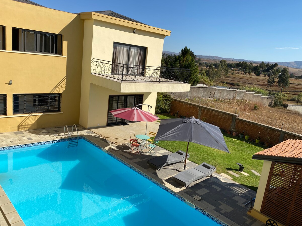 An outdoor area features a large pool surrounded by lounge chairs and umbrellas in varied colors. A grassy space and a stone patio complement the setting, while a two-story building is visible in the background, offering large windows for natural light.