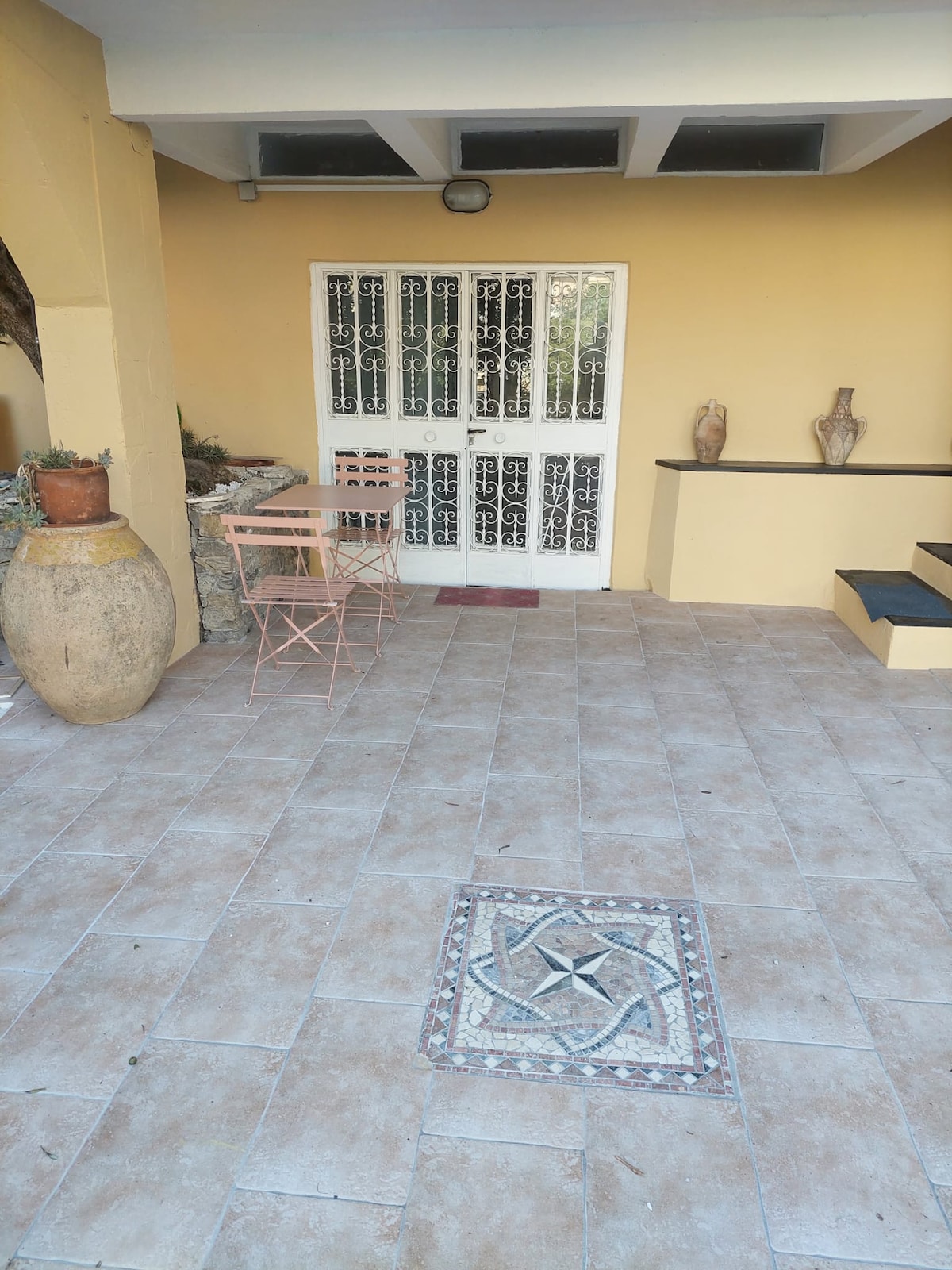 An entrance area features a patterned tile section on the floor, complemented by a small table and two chairs. Decorative pottery can be seen nearby, and a glass door with intricate metalwork provides access to the interior.