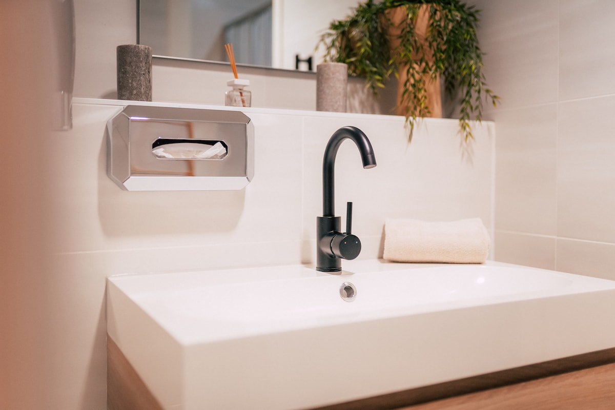 A modern bathroom is depicted, featuring a sleek, white sink with a minimalist faucet. A neatly folded towel rests beside the sink, and decorative items are positioned on the counter. Greenery adds a touch of nature to the space.