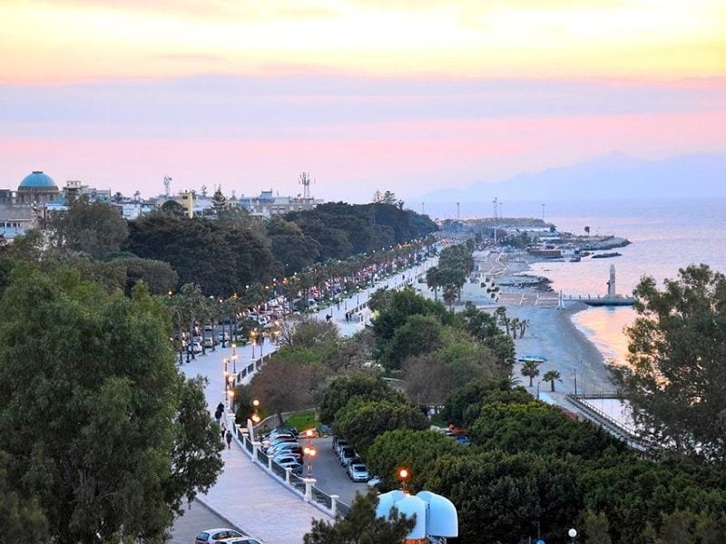 A scenic view of the coastline is captured at sunset, displaying a long promenade lined with trees and streetlights. The beach and calm sea are visible in the distance, while a path is filled with people enjoying the tranquil evening atmosphere.