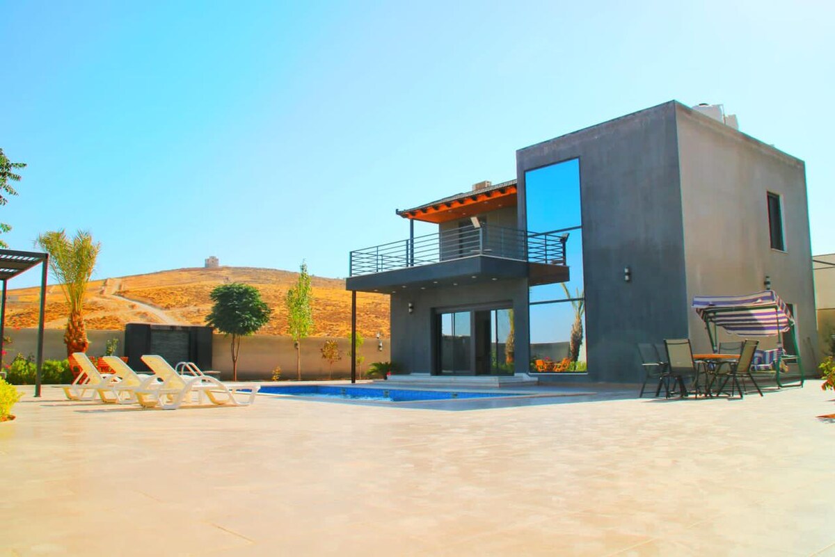 A modern villa is depicted, featuring large glass windows that reflect the surrounding landscape. The outdoor space includes a private pool, lounging chairs, and a dining area. The serene backdrop of a hill enhances the peaceful ambiance of the setting.
