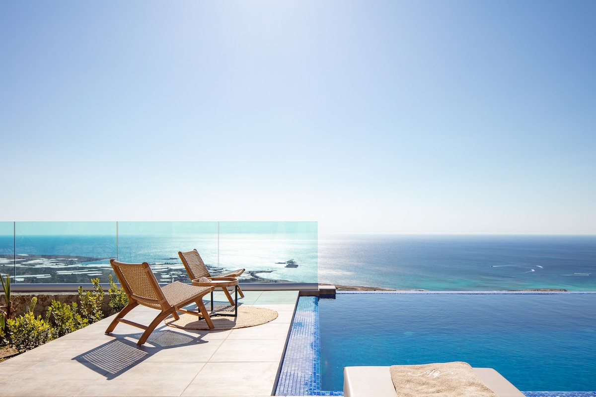 A serene outdoor space features two wooden lounge chairs positioned beside an infinity pool. The pool's edge merges with the stunning view of the Cretan Sea, where the endless blue sky meets tranquil waters, creating a peaceful backdrop.