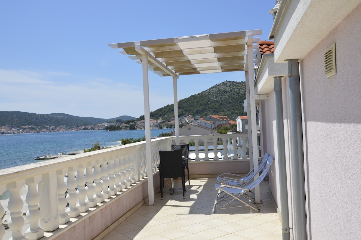 A balcony is shown overlooking the sea, featuring a pergola for shade. Two sun lounges are positioned beside a small table with a chair, providing a comfortable space to enjoy the view of the water and surrounding hills.