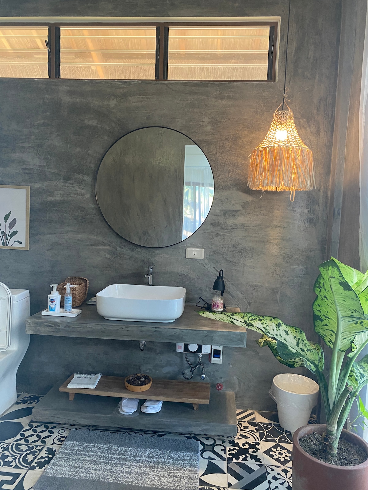 A modern bathroom features a round mirror above a sleek, rectangular sink positioned on a concrete countertop. Decorative items and toiletries are arranged on the shelf below, while a plant adds a touch of greenery. Natural light enters through large windows, enhancing the neutral tones of the space.