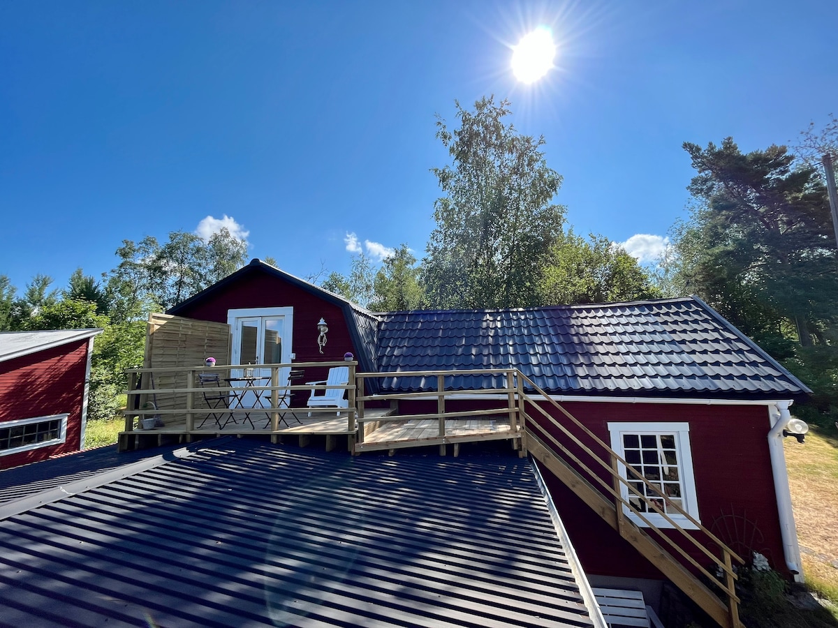 An outdoor deck is seen atop a red building, featuring white chairs positioned for relaxation. Sunlight brightens the scene, casting a warm glow over the roof and surrounding trees.