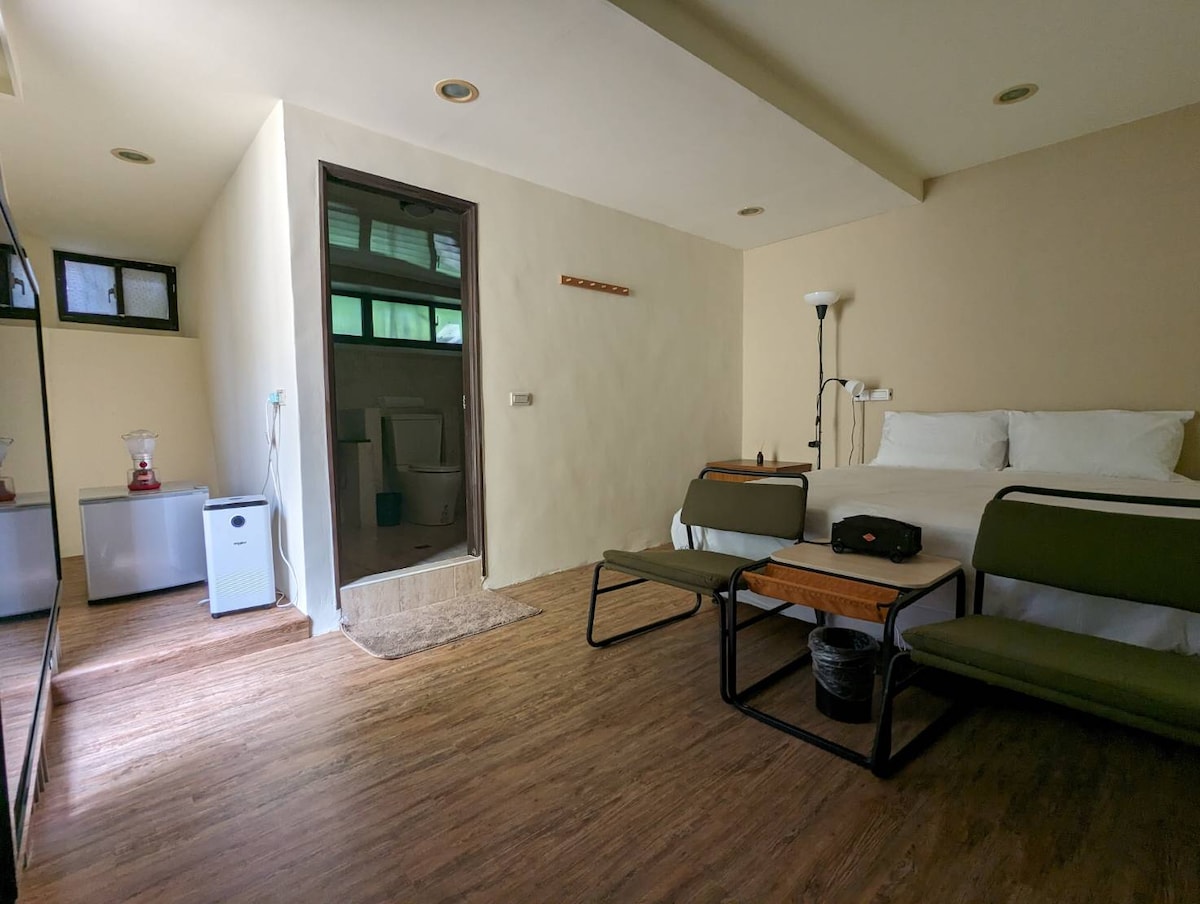 A spacious room features a large bed with crisp white linens. Simple wooden furniture includes two chairs and a small table. A mirrored wardrobe reflects the natural light from the windows. A door leads to a private bathroom, enhancing convenience.