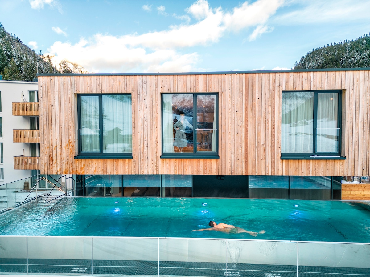 A heated outdoor pool is featured in the foreground, with a swimmer gliding through the water. The building above showcases a modern wooden facade with large windows, offering views of the surrounding landscape and mountains.