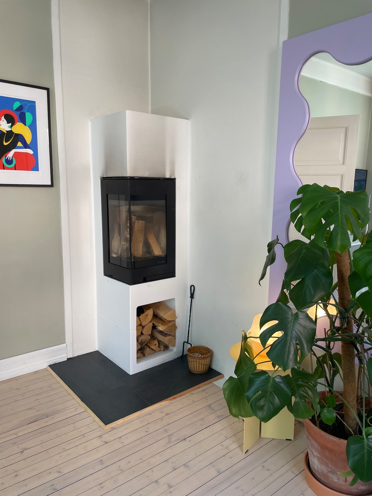 A modern fireplace is positioned in the corner, featuring a glass front and a storage area filled with firewood below. A colorful piece of wall art is hung above it, complemented by a large green plant in a terracotta pot next to a warm light source.