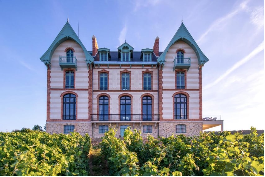 A grand three-story building with distinctive architecture is surrounded by lush vineyards. The structure features numerous large windows, providing ample natural light. Its elegant roof peaks add character while a spacious terrace is visible on the side, offering views of the surrounding landscape.