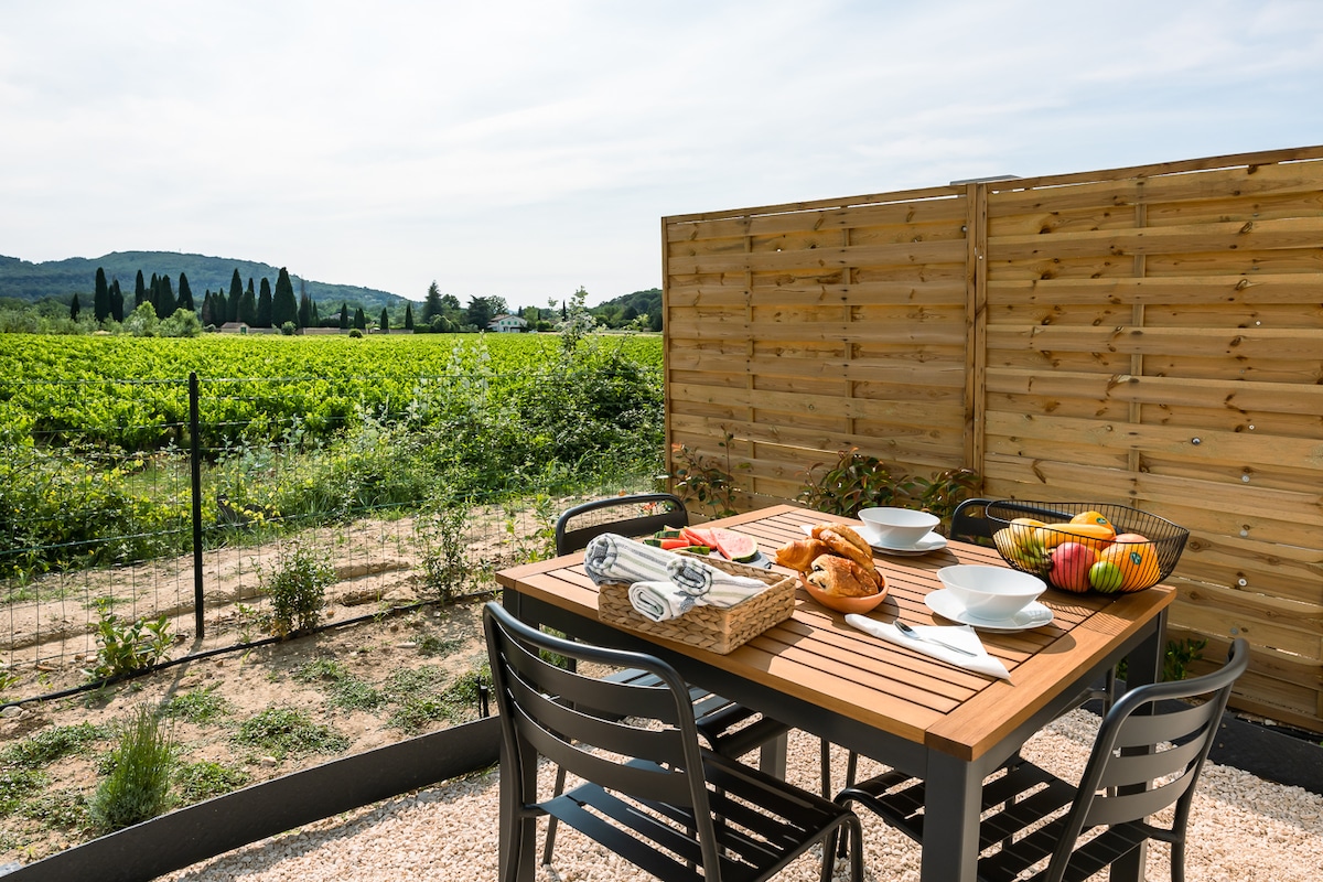 An outdoor dining area is set with a wooden table and black chairs, surrounded by a tranquil vineyard view. A basket of fruit and plates are arranged on the table, while a neatly folded towel adds a touch of preparation for a meal in the natural setting.
