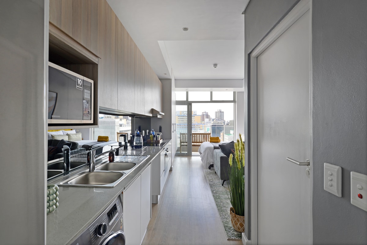 A modern kitchen area is shown, featuring sleek cabinetry and appliances, including a microwave and two sinks. Light floods the space from a large window, illuminating the open layout that connects to a cozy living area and balcony, creating a welcoming ambiance.