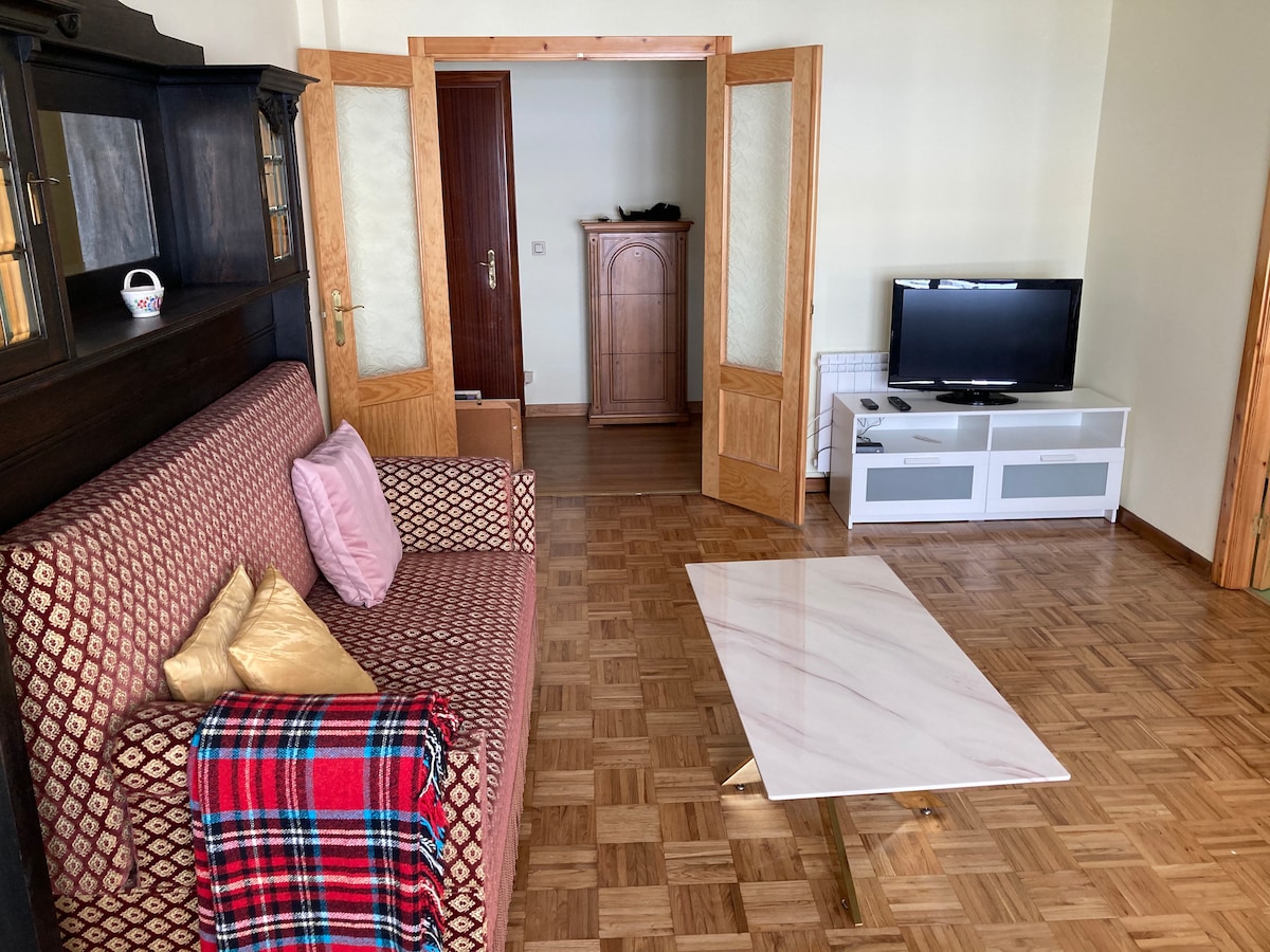 A living room area features a patterned sofa accompanied by a decorative throw and pillows. A marble coffee table sits on a wood-paneled floor. The space includes a TV mounted on a modern white unit, with light entering through partially open doors.