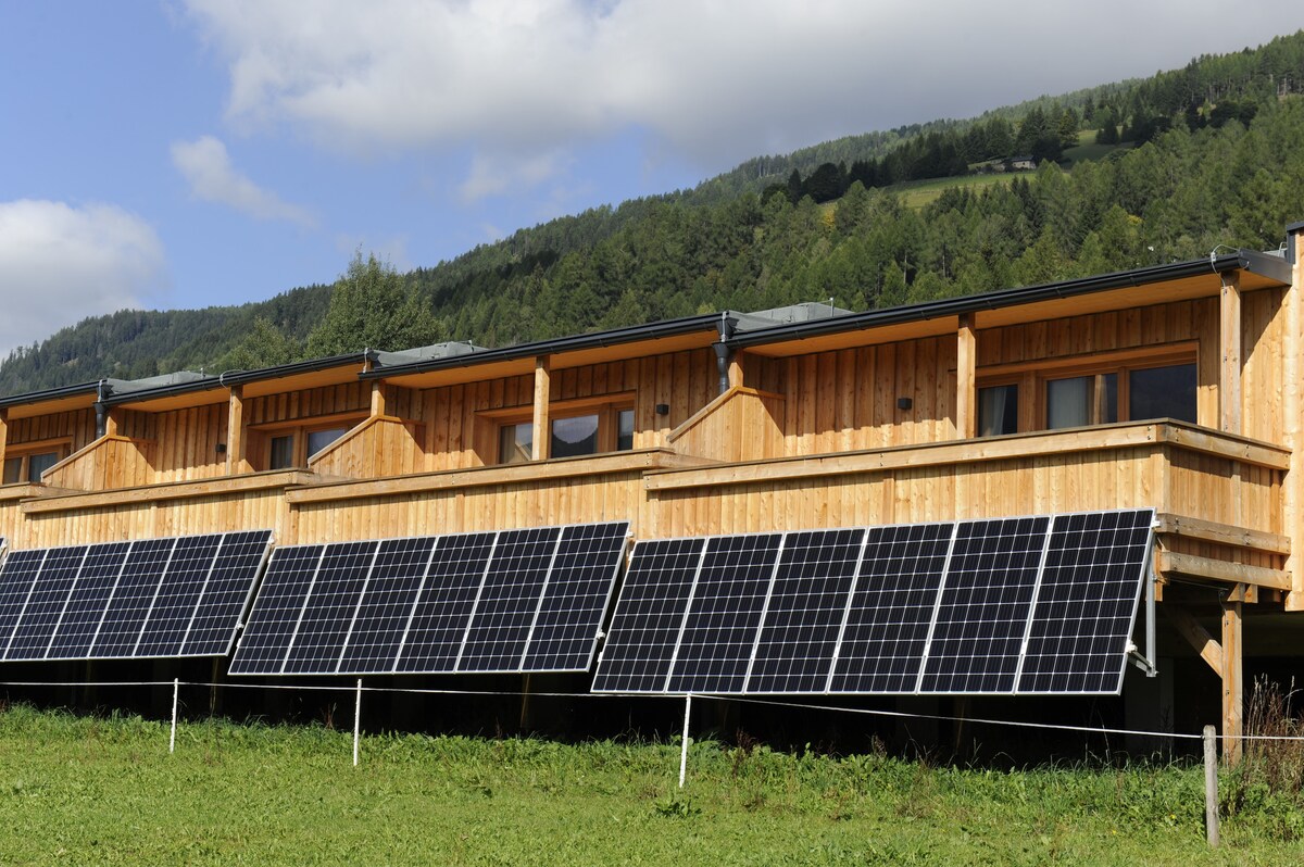 A wooden structure is showcased, featuring multiple large windows that provide natural light. Solar panels are positioned in front, efficiently harnessing energy. A backdrop of green hills under a partly cloudy sky adds to the serene setting.