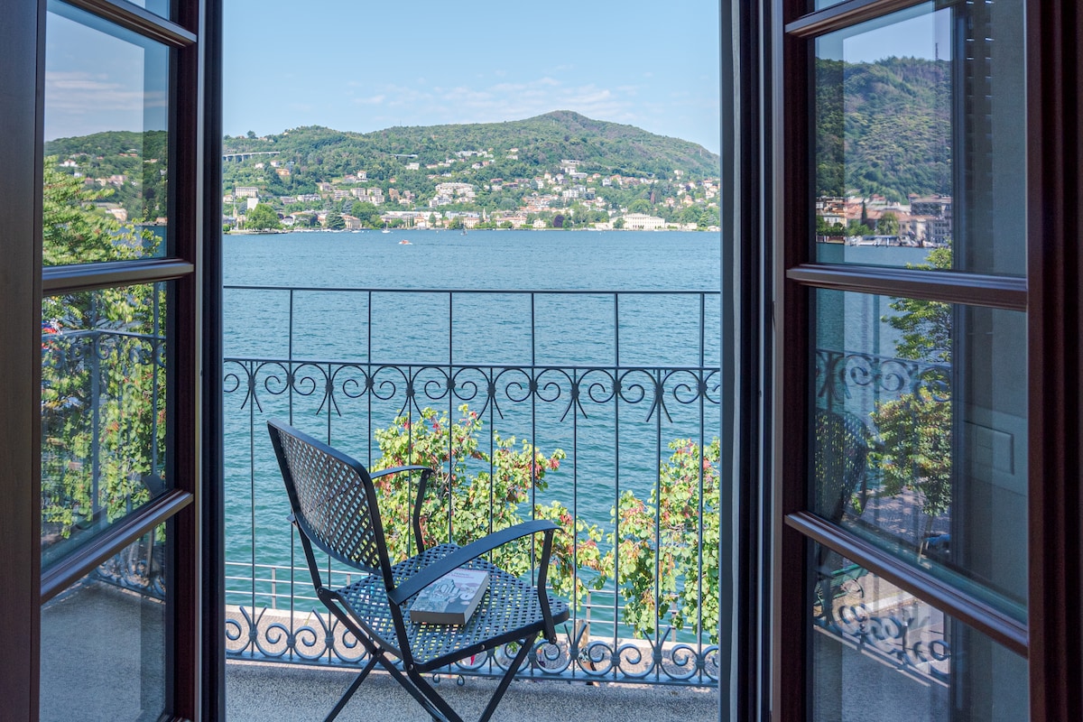 A balcony view is presented, featuring a wrought iron railing overlooking a serene body of water. A single black chair is positioned on the balcony, framed by open double doors. Lush greenery and distant hills contribute to the tranquil setting.