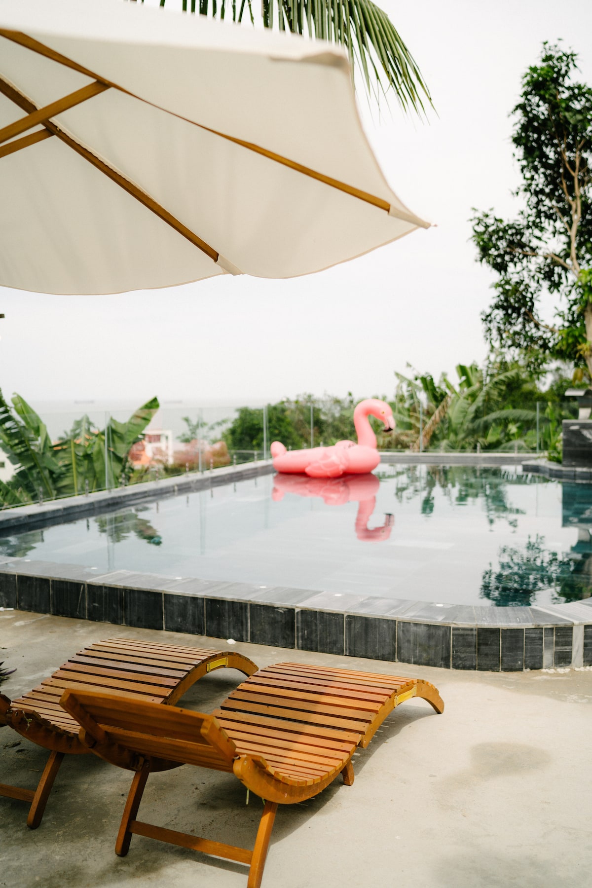 A serene outdoor pool area features a spacious swimming pool surrounded by greenery. A pink inflatable swan floats on the water, while two wooden lounge chairs are positioned nearby, offering a comfortable spot to relax under a large umbrella.