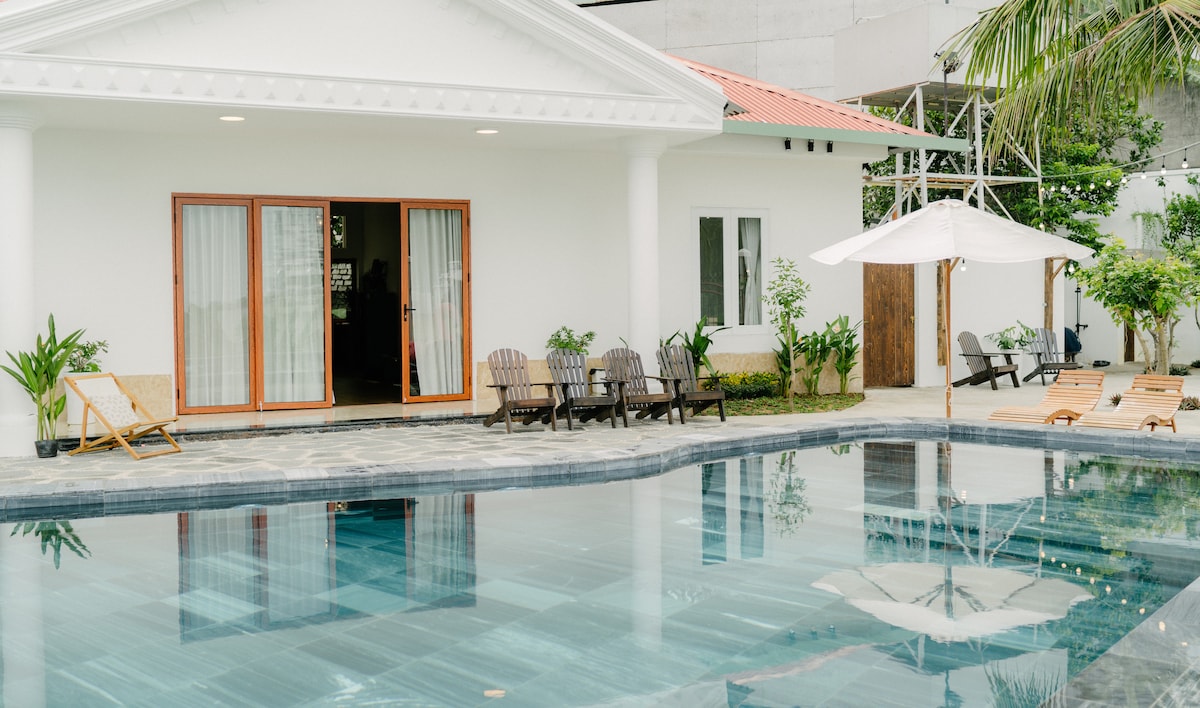 An inviting pool area is showcased with a large, clear swimming pool surrounded by lounge chairs and patio furniture. The villa's exterior features large glass doors leading inside, complemented by lush greenery and a cozy atmosphere.