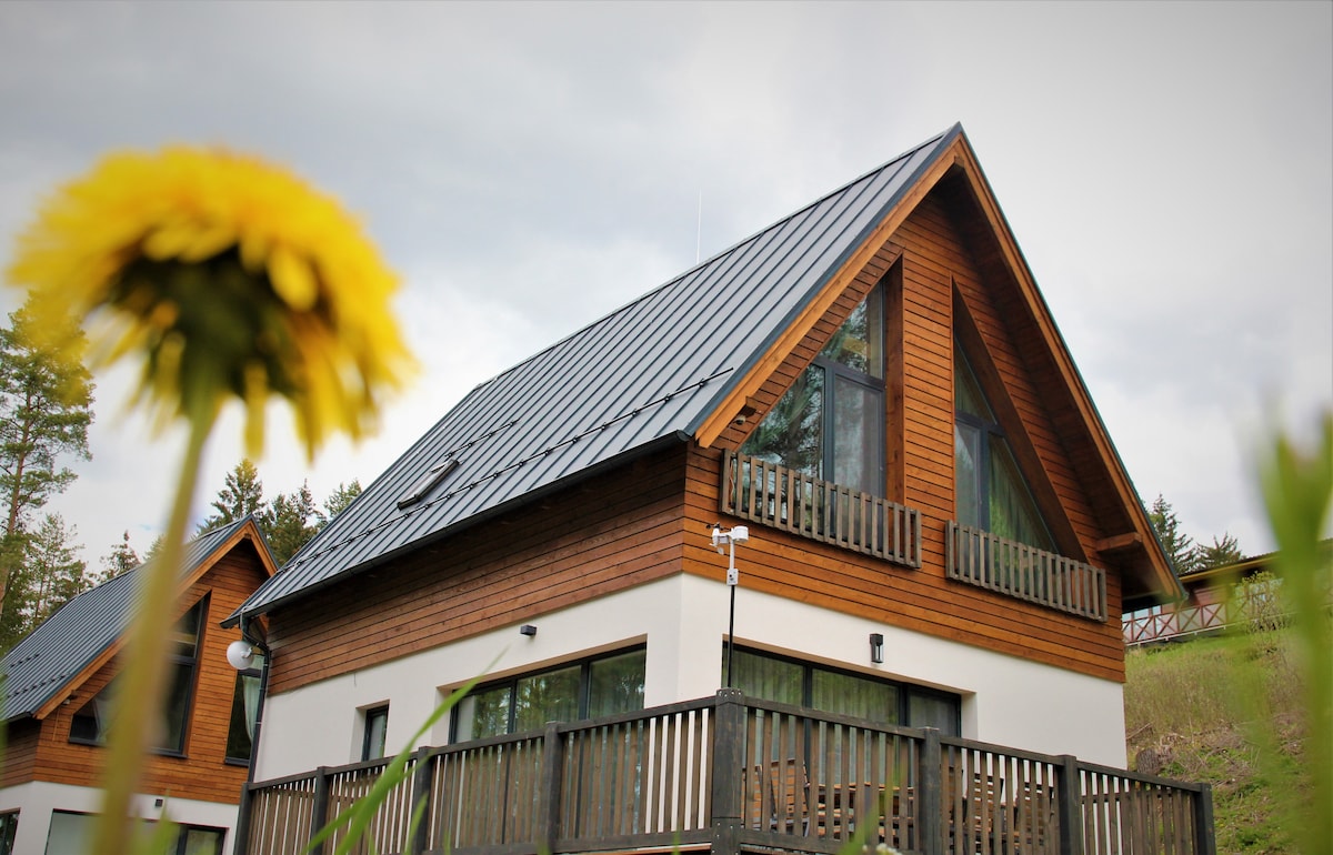 A chalet featuring a sloped roof and large windows is framed by greenery. The wooden exterior blends naturally with the surrounding landscape, while a balcony provides an outdoor seating area. A vibrant yellow flower is visible in the foreground, adding a touch of nature to the scene.
