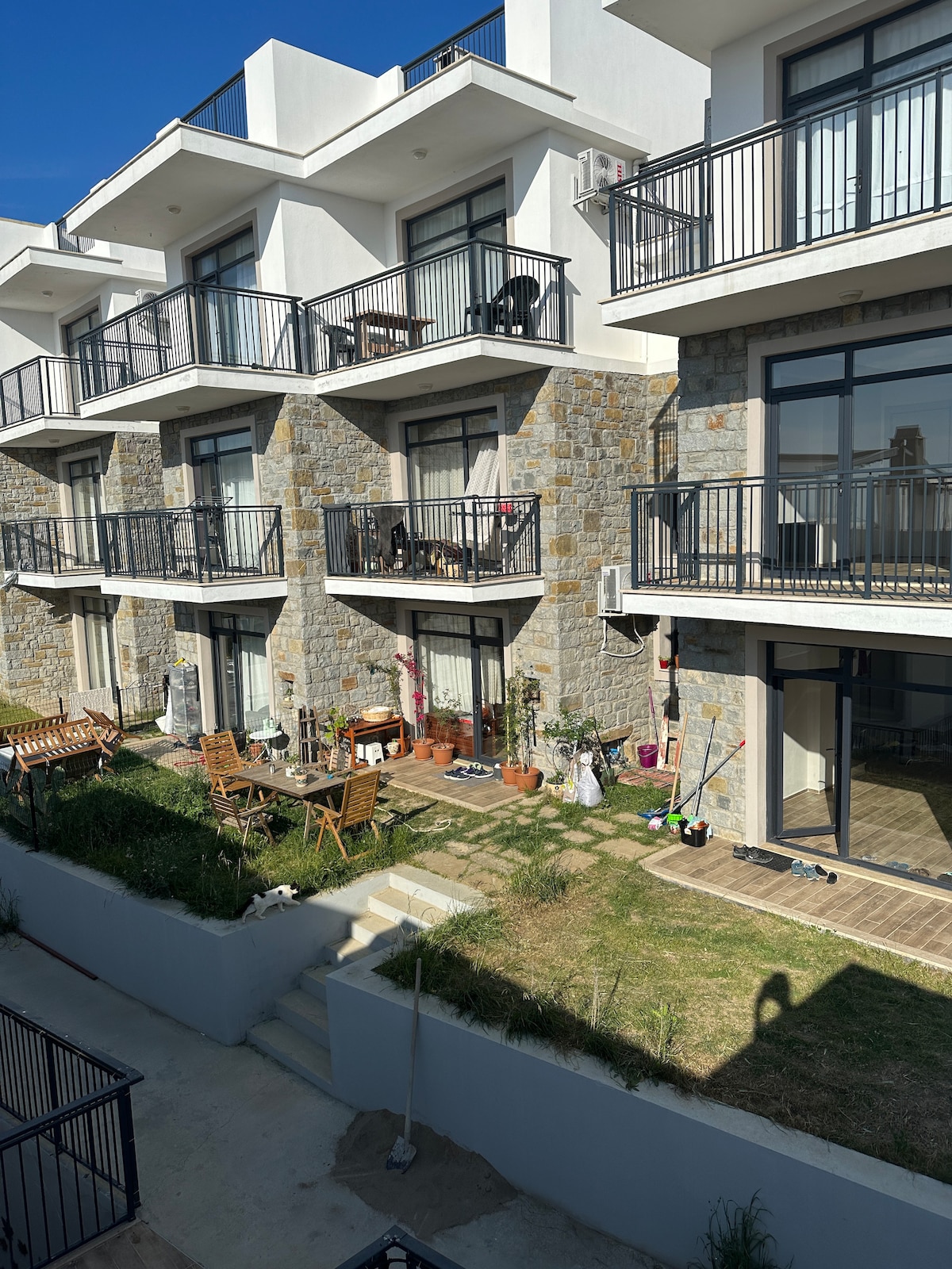 A modern two-story building features stone and white architectural elements. Balconies extend from the upper level, each adorned with seating. A green space below is visible, with outdoor furniture and plantings adding to the inviting atmosphere.