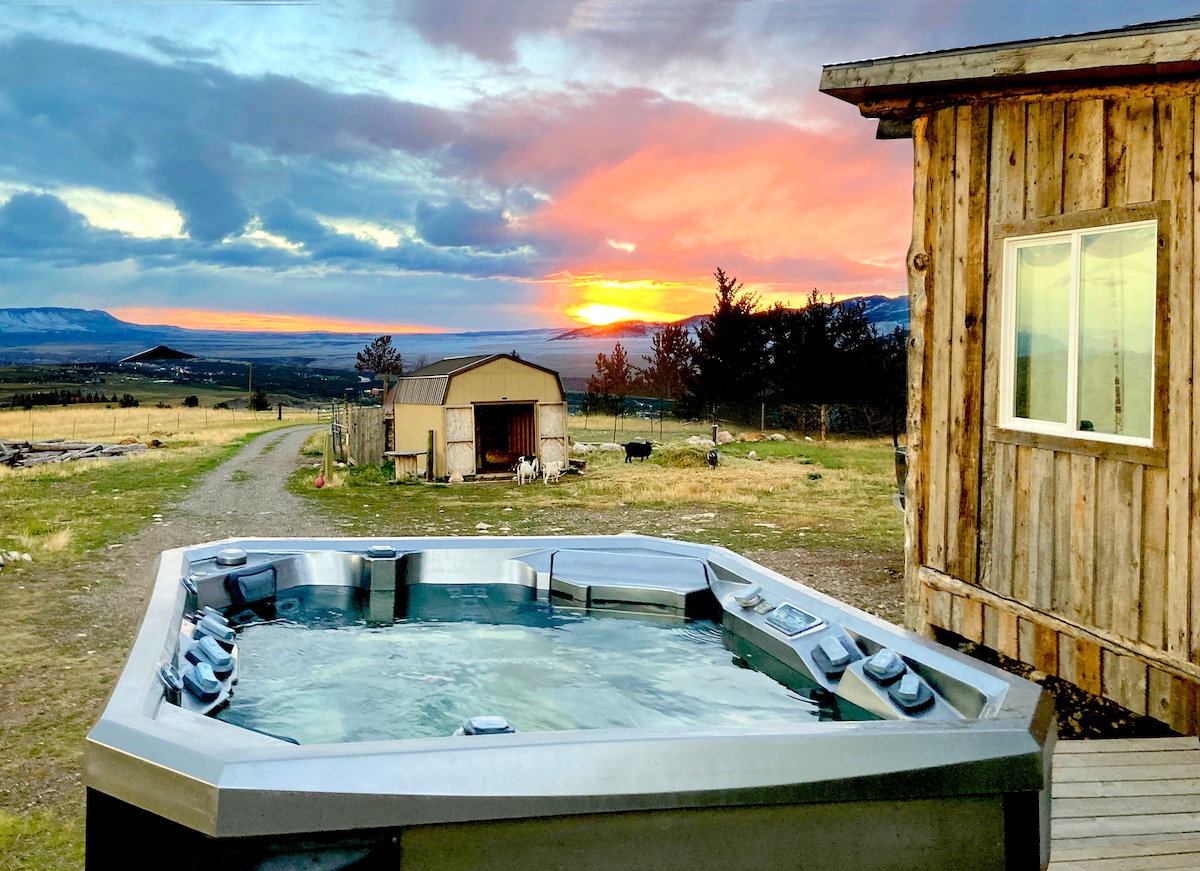 An ozonated jacuzzi is set on a wooden deck, overlooking expansive views of fields and distant mountains under a vibrant sunset. A cozy rustic building and a small barn can be seen nearby, contributing to the serene rural landscape.