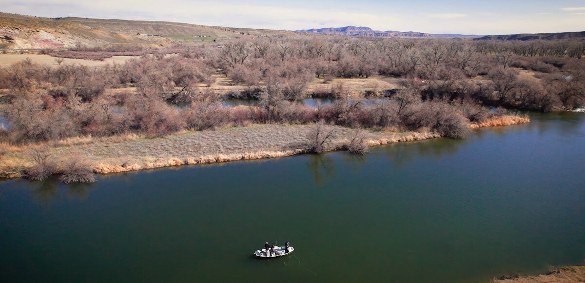A serene river scene features a boat with individuals fishing, surrounded by lush greenery and bare trees along the shore. Calm waters reflect the landscape, creating a peaceful atmosphere. Rolling hills and distant mountains are visible in the background under a clear sky.