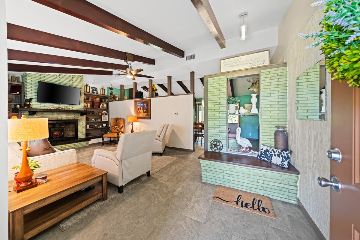 An open living area featuring high wooden beams and a stone fireplace serves as the focal point. Soft seating, including light-colored armchairs, complements a wooden coffee table. A decorative bench with a welcoming ‘hello’ mat adds charm, while shelves display thoughtful decorative items.