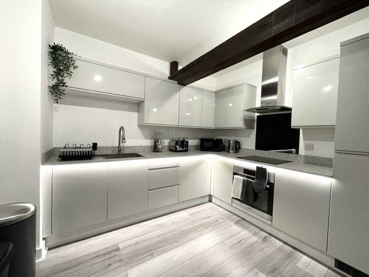 A modern kitchen is equipped with sleek white cabinetry and granite countertops. Stainless steel appliances are integrated, including an oven, microwave, and kettle. Under-cabinet lighting offers a soft glow, and a sink is positioned beneath a wall-mounted shelf with greenery.