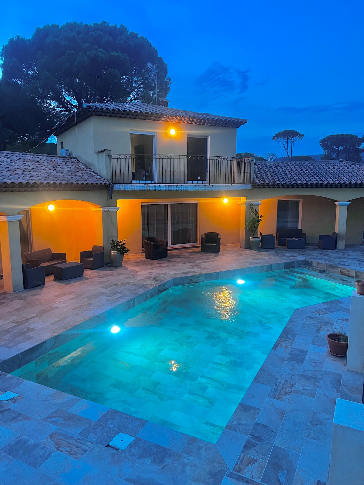 The home is showcased at dusk, highlighting a tranquil swimming pool with illuminated blue water. Soft lighting adds warmth to the exterior, where comfortable seating is arranged on a stone terrace. The second story balcony offers a view of the pool area below.