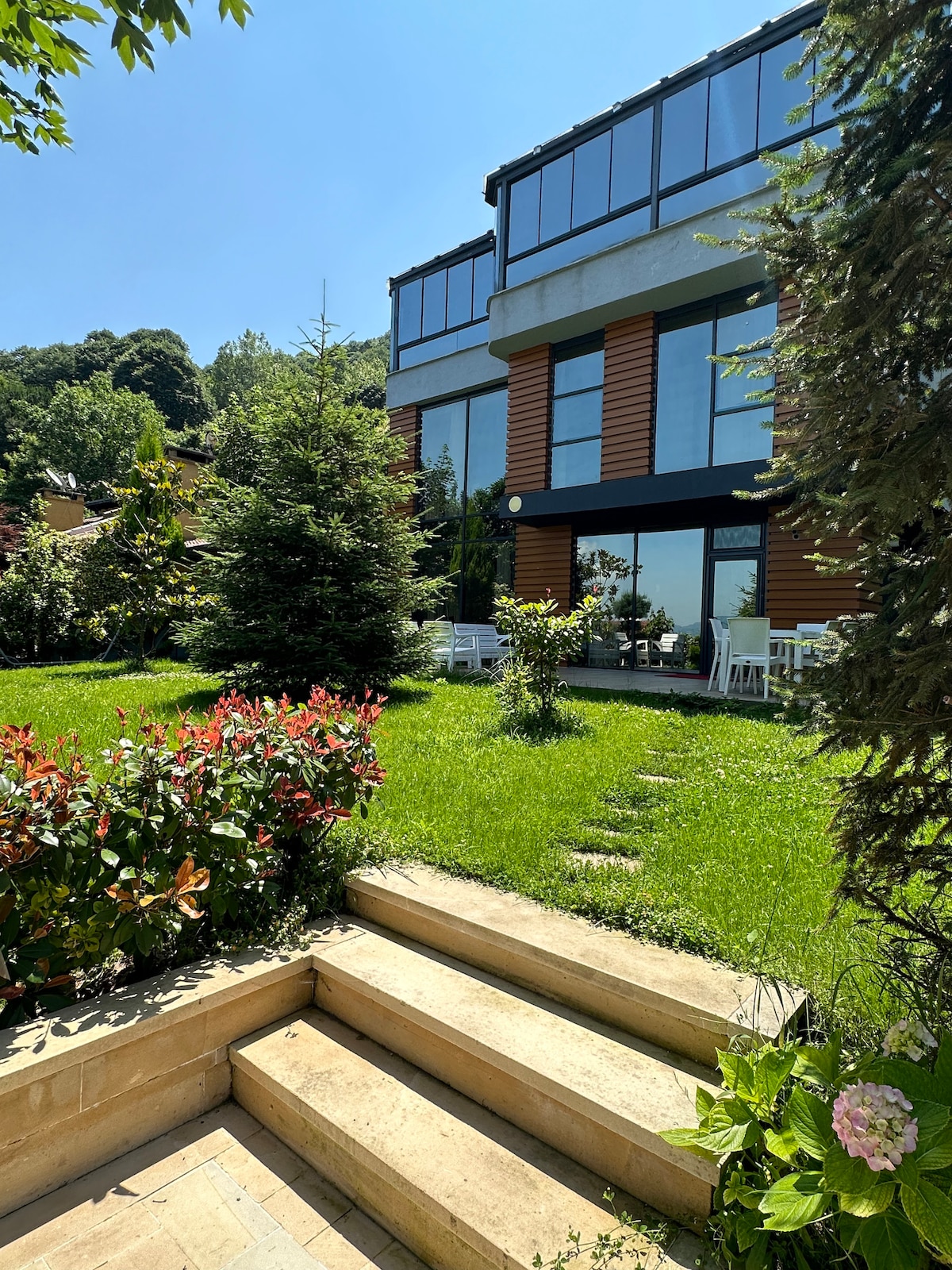 A large modern villa is depicted, featuring expansive windows and a mix of wood and stone siding. Lush greenery surrounds the property, with flowering plants and manicured grass visible. A set of stone steps leads to the entrance, complemented by patio furniture in view.