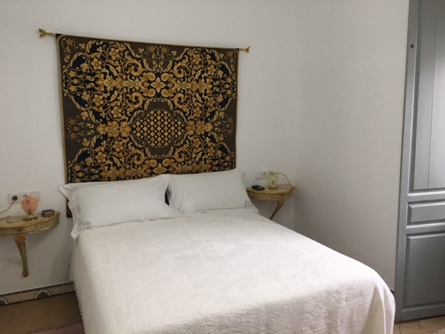 A comfortable sleeping area features a spacious bed covered in a white quilt. Decorative gold-patterned wall art adds a touch of elegance above the bed. Two small nightstands are positioned on either side, each holding a lamp and personal items.