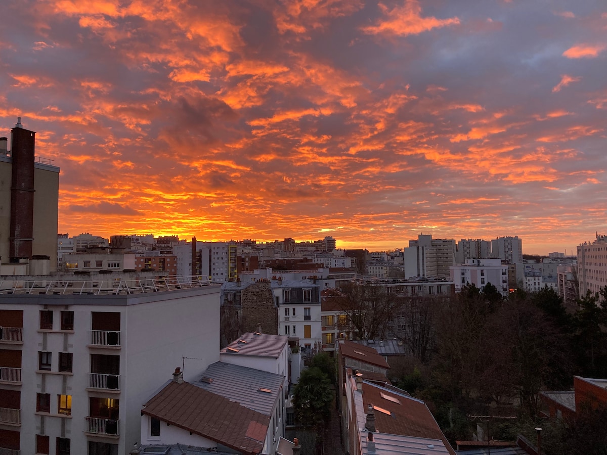A vibrant sunset fills the sky with shades of orange and red, casting a warm glow over a cityscape. Buildings of varying heights are visible, with rooftops and trees in the foreground, creating a tranquil urban scene.