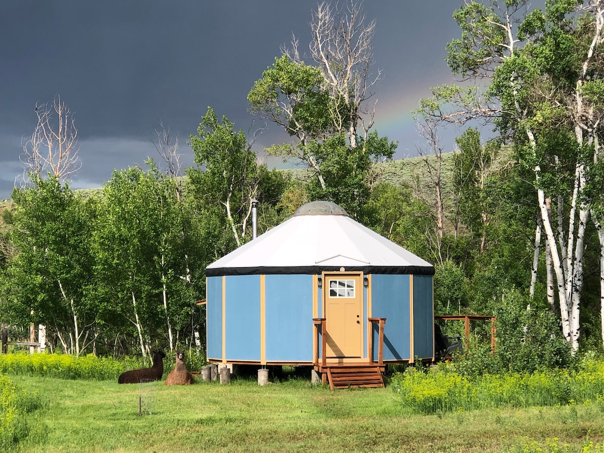 The yurt-cabin is set amidst lush greenery, featuring a circular design with a white and blue exterior. A small wooden deck leads to the entrance, framed by vibrant trees and wildlife, with a rainbow visible in the background.
