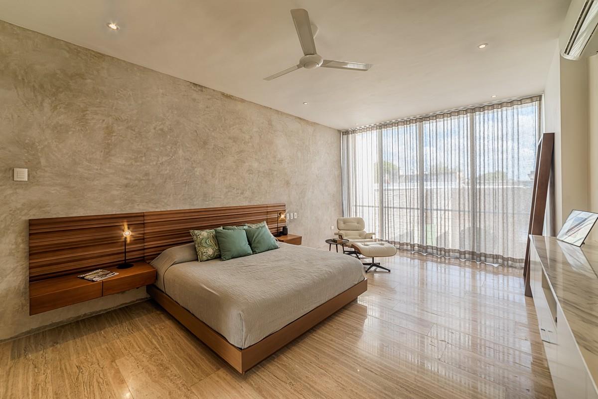 A modern bedroom features a spacious bed with natural wood headboard and soft grey bedding. Natural light floods the room through floor-to-ceiling sheer curtains, casting gentle shadows on the polished marble floor. A comfortable chair is positioned nearby, enhancing the room's serene atmosphere.