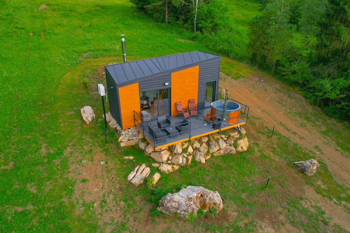 A contemporary cabin is situated on rocky terrain, surrounded by lush greenery. The building features a sleek exterior design with wooden accents. A private hot tub is positioned on a wooden deck, accompanied by seating areas for relaxation. The peaceful natural setting enhances the overall retreat experience.